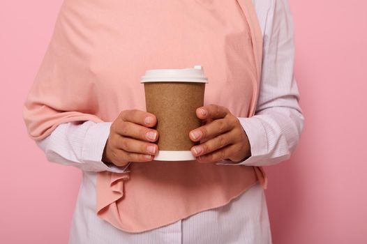 Close-up studio shot of female hands holding a craft takeaway cardboard cup with hot drink. Cropped view of Muslim woman in hijab with disposable paper mug, isolated on pink background with copy space