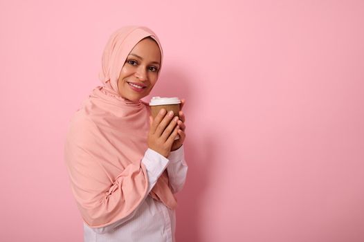 Arabic muslim pretty woman with covered head in pink hijab drinking hot drink, tea or coffee from disposable cardboard takeaway cup, standing three quarters against colored background with copy space