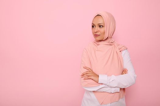 Confident portrait of Arabic Muslim beautiful woman with attractive gaze and covered head with pink hijab, stands three quarters to colored background with copy space and looks away at copy space