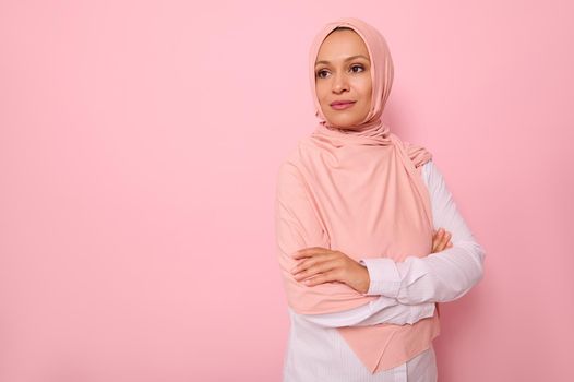 Confident portrait of Arabic Muslim beautiful woman with attractive gaze and look, wearing pink hijab and standing three quarters to colored background with copy space