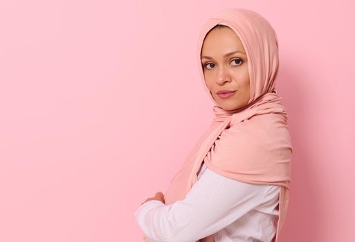 Confident close-up portrait of Arabic Muslim beautiful woman with attractive look and gaze, wearing a pink hijab, looking at camera confidently standing sideways against colored background, copy space