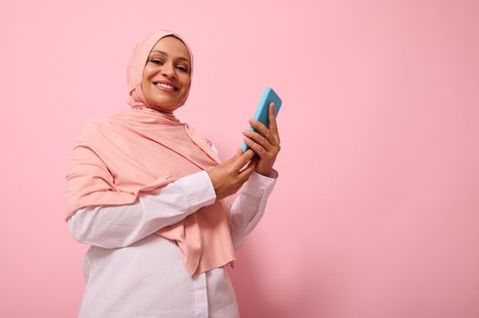 Smiling with toothy smile mature Muslim woman of Middle Eastern ethnicity in colored hijab with smartphone in blue cover on hands, looks at camera, isolated on pink pastel background, copy space