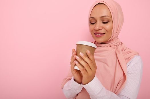 Arabic muslim pretty woman with covered head in pink hijab drinking hot drink, tea or coffee from disposable cardboard takeaway cup, standing three quarters against colored background with copy space
