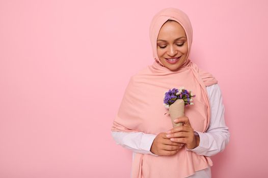 Isolated portrait on colored background with copy space of an attractive charming Muslim Arab woman in pink hijab, looking at a cute bouquet of wildflowers in purple shades, wrapped in craft paper