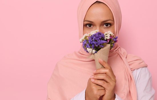 Close-up headshot portrait of young charming Arab Muslim woman in pink hijab with beautiful dark eyes, attractive gaze, looking at camera, covers half of face and mouth with a craft bouquet of flowers