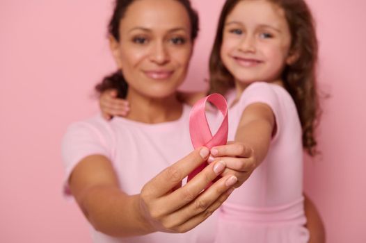 Focus on pink satin ribbon in the hands of two generations of women, mother and daughter posing on pink background, showing support, solidarity to patients and survivors. World Day of fight cancer