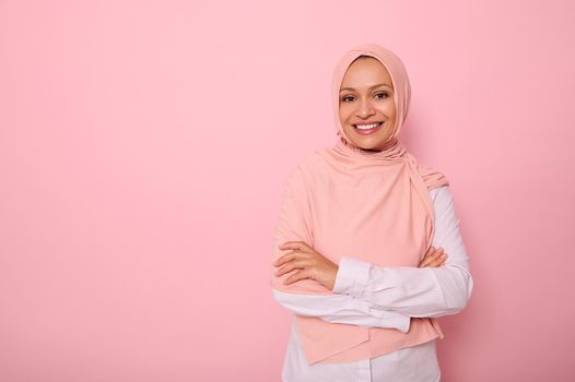 Arab Muslim lady wearing a hijab crossing arms, smiling with beautiful toothy smile, poses on pink background with copy space looking at camera. Concept of successful confident islamic modern woman