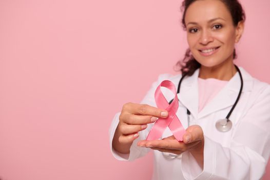 Soft focus on Breast Cancer Awareness Ribbon, in hands of blurred mixed race woman doctor in white medical coat, isolated on pink background, copy space. 1 st October, World Day of fight breast cancer