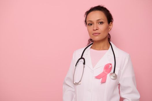 Confident portrait of a mixed race female doctor in medical coat with pink ribbon, and stethoscope around neck, looking at camera, isolated on colored background, copy space. Breast Cancer Day concept