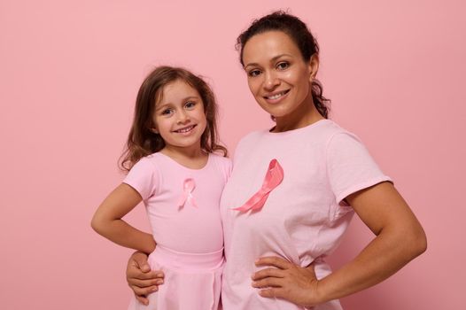 Isolated portrait on colored background with copy space of mixed race woman hugging her daughter, wearing pink clothes and breast cancer awareness ribbon showing solidarity with cancer survivors