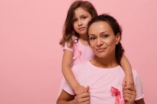 Woman and girl in pink attire with Breast Cancer Awareness ribbon, daughter hugging her mother, holding by hands, looking at camera, supporting cancer patients. Pink background with copy space.