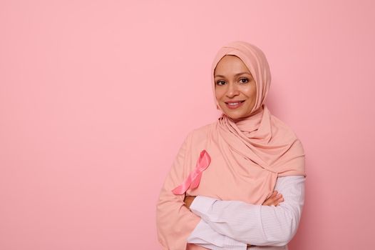 Isolated portrait of smiling Arab Muslim woman wearing pink hijab with pink satin ribbon in symbol of International Day of Cancer Awareness. Oncological diseases concept. Copy space on pink background