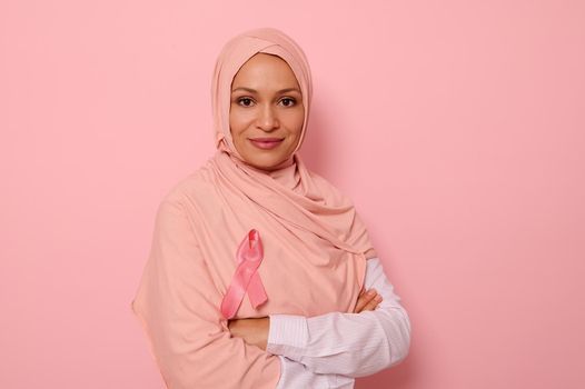 Confident portrait of a friendly Arab Muslim woman wearing a hijab and pink satin ribbon showing her support for cancer patients and survivors. Women's health care. World Breast Cancer Awareness Day.