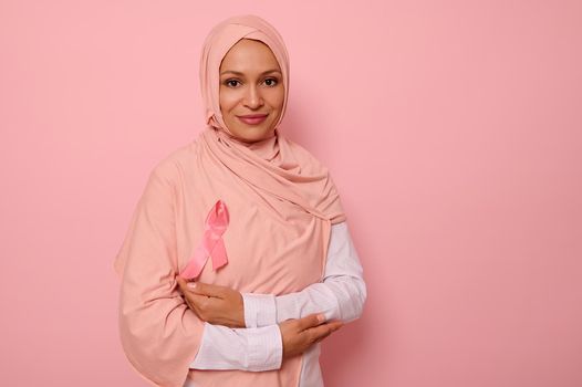Gorgeous Arab Muslim woman with covered head in hijab, stands on pink background with crossed arms on chest and wearing pink ribbon, symbol of Breast Cancer Awareness Day. Medical concept, copy space
