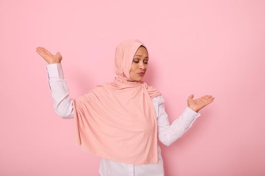 Waist-length portrait of beautiful mysterious Muslim woman wearing a headscarf hijab, showing something on her palms isolated on colored pink background with copy space for advertising or promotion