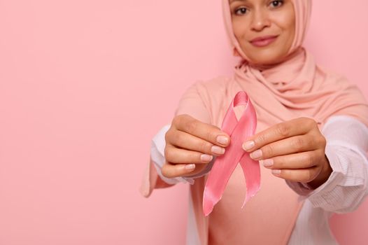 World Cancer Day concept. Focus on pink Breast Cancer Awareness ribbon in the manicured hands of blurred beautiful friendly Arab Muslim woman in pink hijab headscarf on pink background with copy space