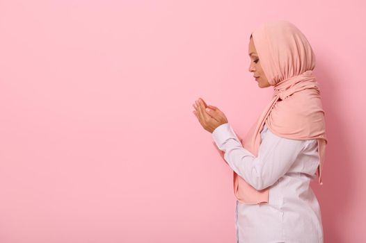 Side portrait of a beautiful and serene Muslim Arab woman in pink hijab and strict outfit with palms folded together at face level performing namaz, isolated on colored background with space for text