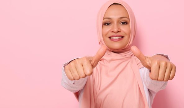 Soft focus on hands of a blurred smiling with toothy smile Arab Muslim woman in pink hijab, showing thumbs up to the camera, isolated over colored background with space for text