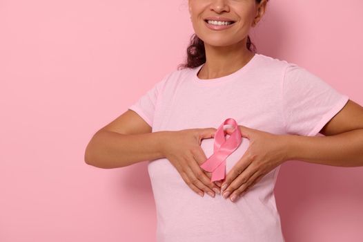 Cropped portrait on colored background of smiling woman in pink t-shirt, putting hands on chest in shape of heart with a pink satin ribbon in the center. World Cancer Awareness Day, fighting cancer