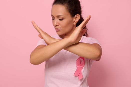 Pretty woman wearing pink t-shirt with breast cancer ribbon looking down on pink background, arms crossed in front of her. International Day of fight Cancer disease, World Breast Cancer Awareness Day