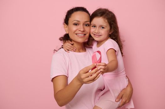 Two generations of women, woman and girl, mother and daughter posing against pink background, holding a pink ribbon, showing support and solidarity to patients and cancer survivors. Medical awareness