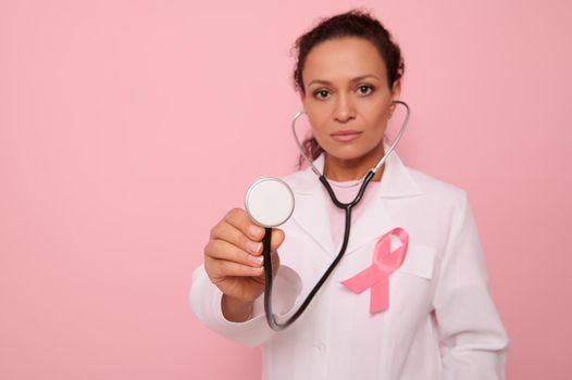 Focus on phonendoscope in the hands of beautiful mixed race Hispanic doctor in medical coat with pink satin ribbon, symbol of Global Breast Cancer awareness Day, Pink October . Woman's health concept