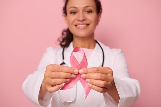 Soft focus on Breast Cancer Awareness Pink Ribbon, in hands of blurred mixed race woman doctor in white medical coat, isolated on colored background. 1 st October, World Day of fight breast cancer