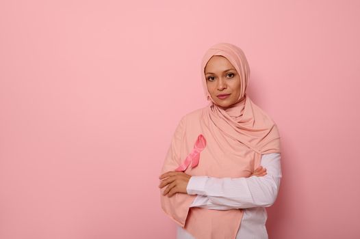 Confident portrait of gorgeous Middle Eastern ethnicity Arab woman, wearing pink hijab with pink Breast Cancer Awareness ribbon, looking at camera, posing over pink background with copy space