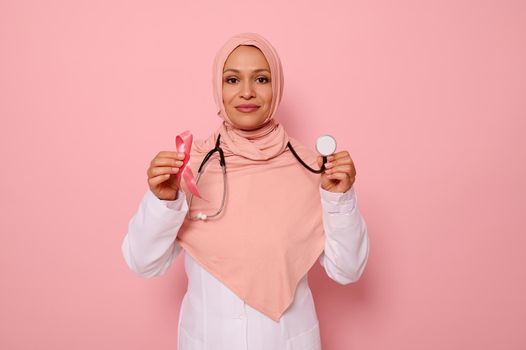 Portrait of Muslim Arab woman doctor with covered head in pink hijab, posing on colored background with a pink ribbon and stethoscope in hands, supporting cancer patients and survivors, 1 st October