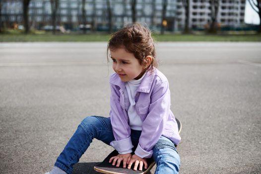 Adorable female child wearing blue jeans and purple jacket sitting on wooden skateboard against blurry buildings background