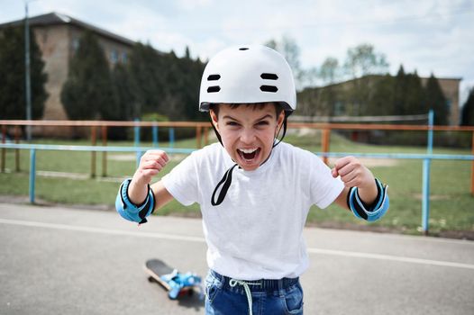 Boy skateboarder in safety helmet screaming and showing fists looking at camera, standing against the background of a playground