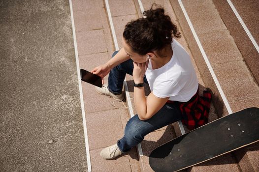 High angle view of a young woman in casual outfit with skateboard sitting on steps and holding a mobile phone