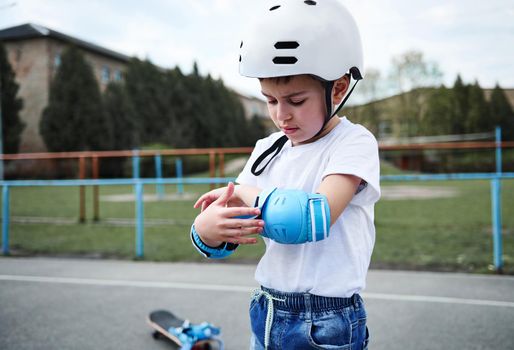 Portraot of a handsome boy skateboarder wearing safety helmet putting on protective armrests before skating. Concept of leisure activity, sport, extreme, hobby.