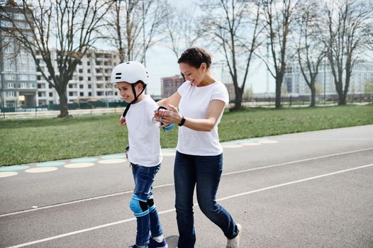 Cute mom stands behind her son in a protective helmet and equipment and helps him to perform skateboarding on the street