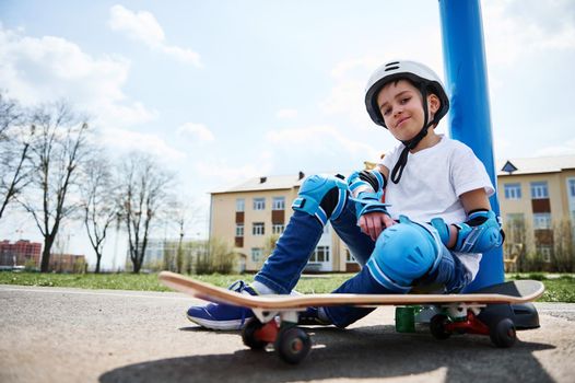 Bottom view of smiling boy in protective gear of skateboarder sitting on skateboard and looking at camera against the background of a yellow building