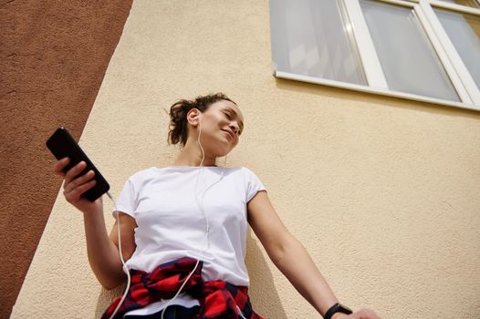 Young woman enjoying listening to music with closed eyes while leaning against a colored wall