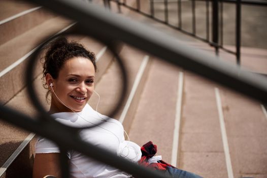 Young mixed race woman wearing headphones smiling looking camera through railing. Closeup