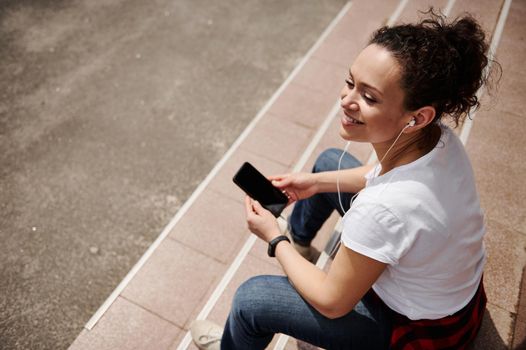Smiling woman with headphones holding a mobile phone and looking away while sitting on steps. High angle view