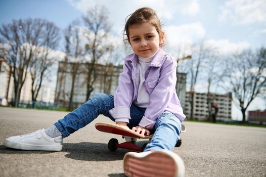 Cute female child posing to the camera while having rest sitting on a wooden skateboard on the playground against the background of blurry buildings