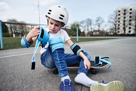 Handsome boy skateboarder in safety helmet and gear sitting on wooden skateboard on playground asphalt