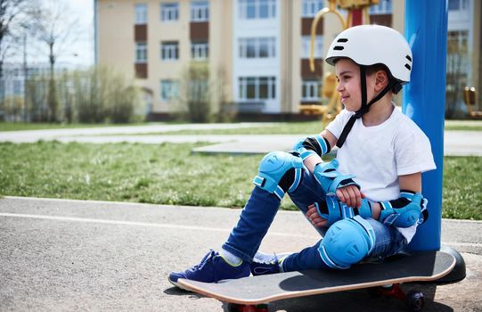 Cute schoolboy relaxed sitting on skateboard against yellow building background