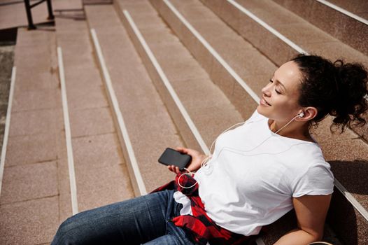 Young beautiful woman leaning on the steps, holding a smartphone in her hands and listening to music on headphones