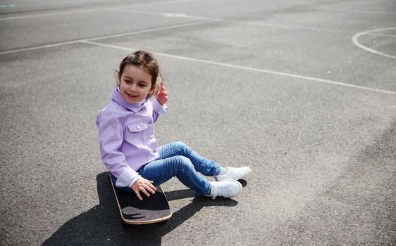 Cute little girl sitting on a skateboard on a sports playground on a beautiful sunny day