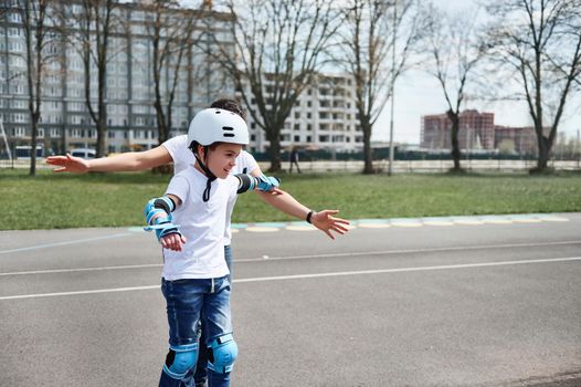 Adorable boy in a skateboard helmet and gear is riding a skateboard on the street against the background of a tall building