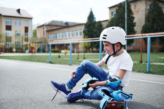 Side view of a sporty boy in safety helmet sitting on a wooden skateboard and putting on protective knee pads