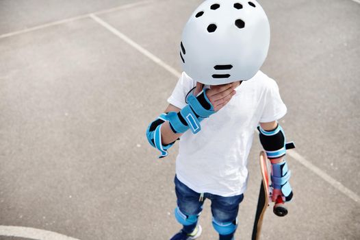 High angle view of a sad boy in protective helmet and gears holding a skateboard, looking down and wiping tears from his eyes, standing on the playground outdoors