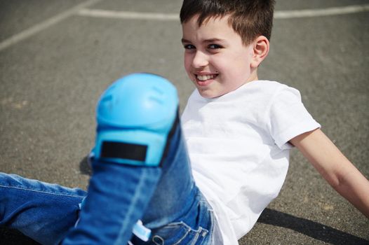 Close-up portrait of a beautiful child in a white t-shirt and protective knee pads sitting on the asphalt outdoors