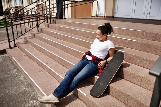 Attractive young African American woman in casual clothing posing on stairs with skateboard and enjoying sunny day