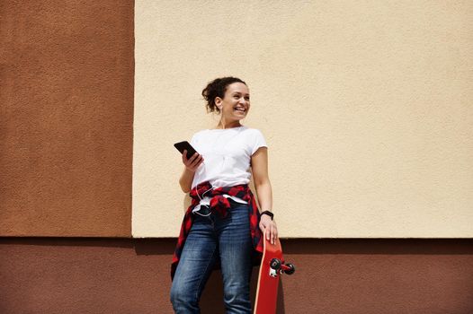 Smiling young woman skateboarder with wooden red skateboard holding smartphone enjoying listening to music while posing outdoors near wall
