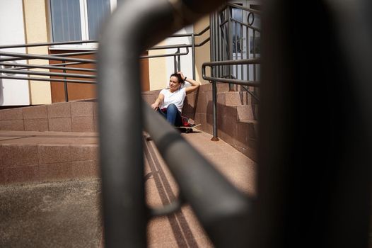 View through the railing of a young woman sitting on skateboard and having rest on a beautiful warm and sunny summer day